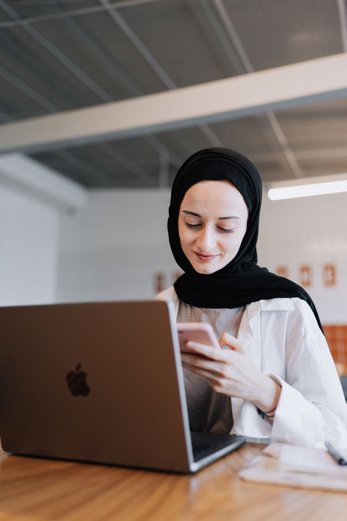 services-04 Smiling woman with hijab using smartphone while working on a laptop indoors.